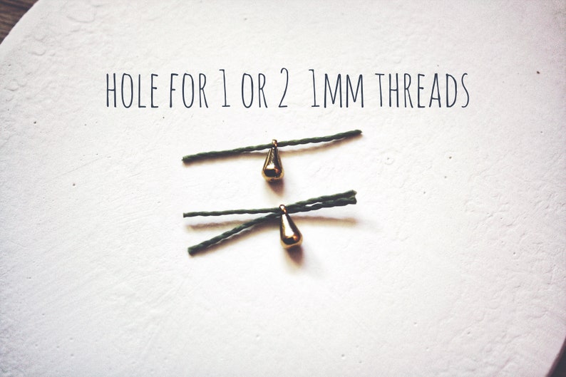 May include: Two gold teardrop-shaped charms with a hole for 1 or 2 mm threads. The charms are hanging from green string.