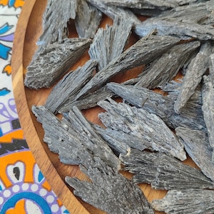 May include: A collection of rough, dark gray kyanite blades arranged on a round wooden tray. The kyanite pieces have a layered, textured appearance, with natural variations in color and shape. The tray sits on a colorful patterned surface.