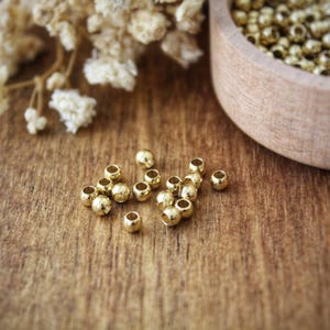 Pode incluir: Close-up de pequenas contas brilhantes douradas espalhadas sobre uma superfície de madeira. Uma tigela de madeira cheia de contas semelhantes está no fundo, juntamente com flores brancas secas. Ideal para a criação de joias.