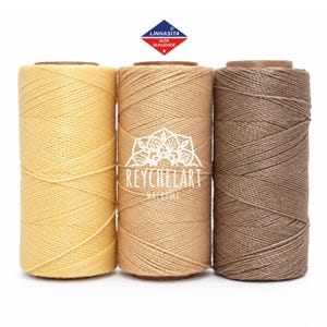 Puede incluir: Tres bobinas cilíndricas de cordón en amarillo, beige y marrón. La bobina central presenta un logotipo blanco con el texto REYCHELART MACRAME. Sobre las bobinas hay un logotipo en forma de rombo azul y rojo con el texto LINHASITA ALTA QUALIDADE.