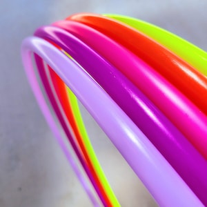 May include: A stack of five smooth, circular plastic hoops in vibrant neon colors. The hoops are arranged in a nested, angled formation, displaying a sequence of light purple, magenta, bright pink, orange, and lime green hues with a glossy finish.