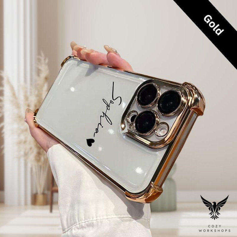 May include: A gold-colored phone case with a clear back. The case has a gold frame and camera surround. The name "Sophia" is written in black script with a small heart. The word "Gold" is printed on a black diagonal banner.