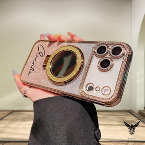 May include: A decorative phone case with a glittery pink and silver design. It features a gold ring holder and a section with three camera lenses and a decorative border of small, sparkling gems. The name "Quentin" is written on the case.
