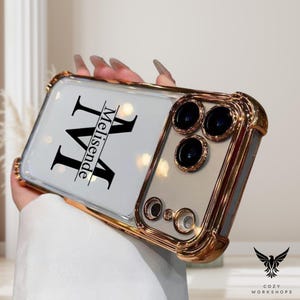 May include: A gold-coloured phone case with a clear back and a white insert featuring a large black letter "M" and the name "Melisende". The case has a gold frame and a camera module with three black lenses.