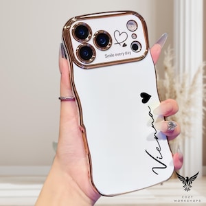 May include: White phone case with a gold-colored frame. The case has a camera bump with three lenses and a heart design. The text "Smile every day" and the name "Vienna" are printed on the case.