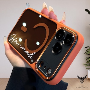 May include: Orange phone case with a brown smiley face design and the name "Hannah" in white script. The case has a gold-colored trim and a black camera module with three lenses. The case is from Cozy Workshops.