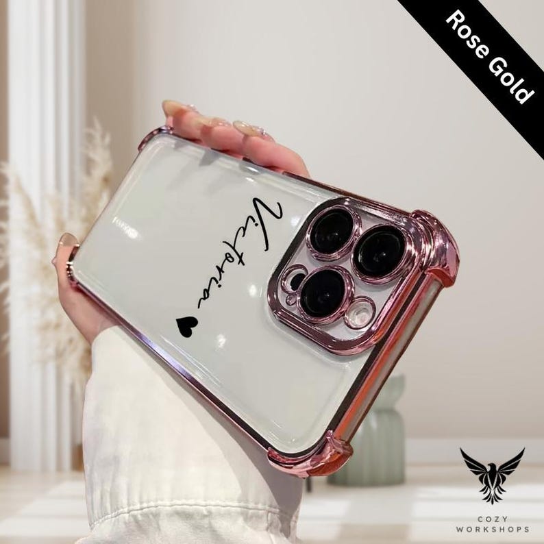 May include: A clear phone case with rose gold trim, featuring the name "Victoria" in black script with a small heart. The case has a glossy white back and a protective bumper design. The words "Rose Gold" are printed on a black diagonal banner.
