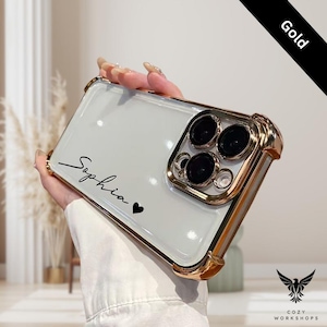 May include: A gold-trimmed phone case with a clear back. The case has the name "Sophia" and a heart symbol printed on it. The case has a gold border and a gold camera lens protector.