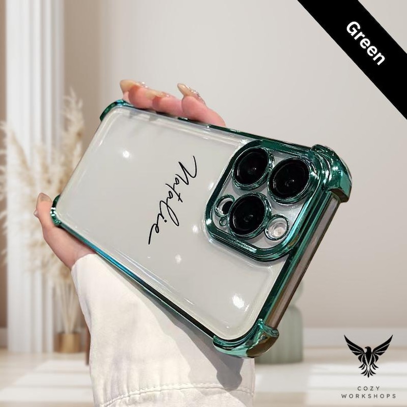 May include: A clear phone case with a green metallic frame and camera surround. The name "Natalie" is written in cursive on the case. The case has a protective design and features the "Cozy Workshops" logo.