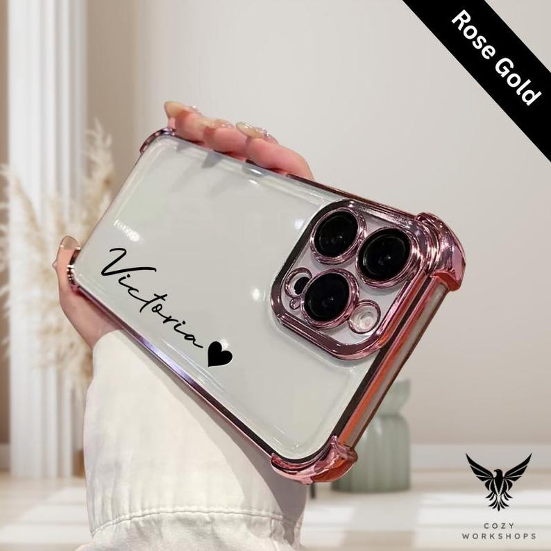 May include: A clear phone case with a rose gold bumper. The case has the name "Victoria" in black script with a black heart. The phone case has a clear back and a rose gold camera ring.
