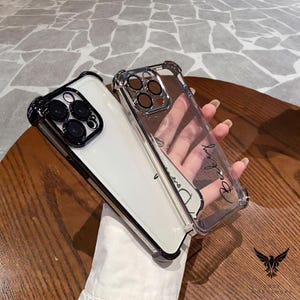 May include: Two clear phone cases with black trim. One case is on a white phone, the other is empty. The empty case has the word "Beauty" written in cursive. The cases have a protective design.