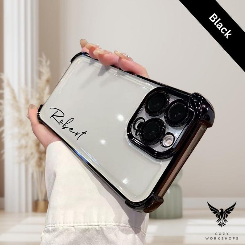 May include: A clear phone case with a black border and camera protector. The case has the name "Robert" in script. The top right corner has a black banner that says "Black". The bottom right corner has a logo that says "Cozy Workshops".