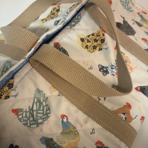 May include: A beige tote bag with a repeating pattern of colorful chickens on a white background. The bag has beige woven handles and a blue interior lining. The chickens are in various colors and patterns, with red combs and yellow beaks.