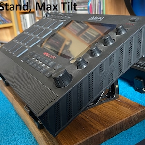 May include: Black wooden stand with a black Akai MPC Live music production sampler tilted at a maximum angle. The stand is on a blue surface.