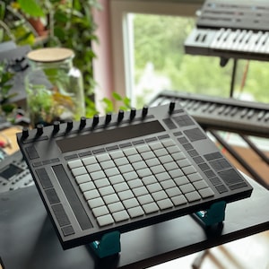May include: A black electronic music production controller with a grid of white square pads and various knobs and buttons. The controller is supported by two teal-colored stands. Other music equipment is visible in the background.