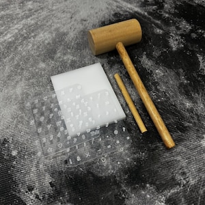 May include: A craft kit featuring a wooden mallet, a small wooden dowel, and clear acrylic letter and number stamps. The stamps include the alphabet, numbers, and symbols. A white rectangular block is also included. The items are arranged on a textured surface.