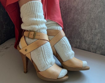 May include: Cream-colored knitted leg warmers paired with light brown leather high-heeled sandals. The sandals feature a buckle closure and a wooden heel. The outfit is set against a red garment and a neutral background.