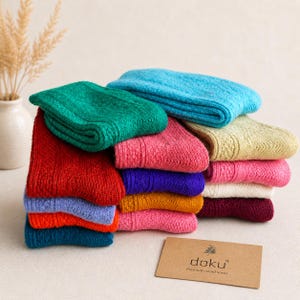 Puede incluir: Una pila de calcetines de lana coloridos en tonos turquesa, verde, rojo, rosa, morado y amarillo. Los calcetines están doblados y cuidadosamente dispuestos, mostrando su diseño de punto texturizado. Una pequeña tarjeta con el texto "doku premium wool socks" es visible.