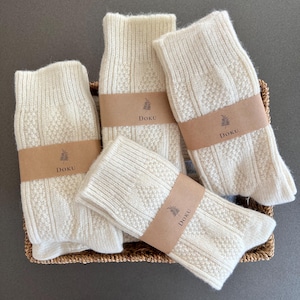 Puede incluir: Cuatro pares de calcetines de punto color blanco roto con un patrón texturizado de trenzas y rombos. Cada par está sujeto con una banda de papel marrón impresa con un icono de árbol y el texto DOKU. Los calcetines están en una cesta de mimbre.