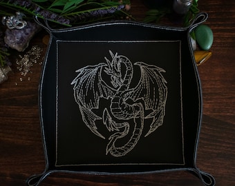 May include: Black square tray with a silver embroidered dragon design. The tray has a stitched border and loop handles. The dragon is centered on the tray, with its wings spread. The tray is on a dark wooden surface.