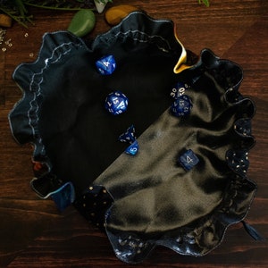 May include: A black satin dice bag with a ruffled edge, holding several blue and white dice. The bag has a contrasting gold dotted pattern on the trim. Perfect for tabletop gaming.