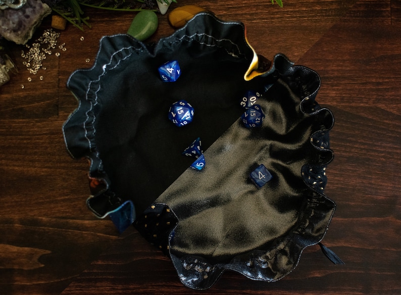 May include: A black satin dice bag with a ruffled edge, holding several blue and white dice. The bag has a contrasting gold dotted pattern on the trim. Perfect for tabletop gaming.