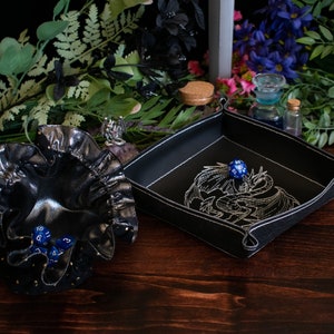 May include: A black leather dice tray with white stitching and a dragon embroidery, next to a black drawstring dice bag. Several blue dice are visible. The items are on a dark wooden surface with floral accents.