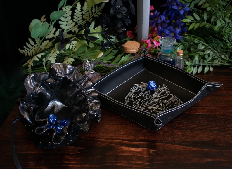 May include: A black leather dice tray with white stitching and a dragon embroidery, next to a black drawstring dice bag. Several blue dice are visible. The items are on a dark wooden surface with floral accents.