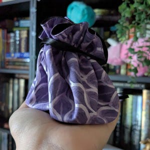 May include: A small, purple drawstring bag with a white, wave-like pattern. The bag is held in a hand and has a black ribbon closure. The background includes a bookshelf and some decorative items.