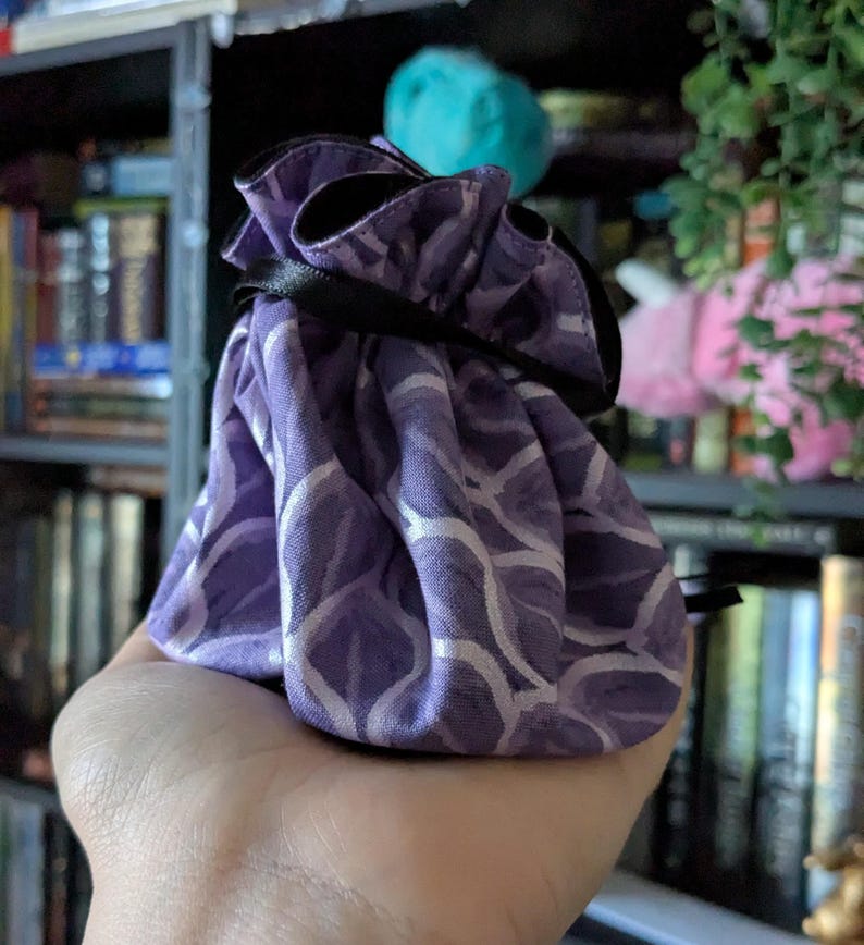 May include: A small, purple drawstring bag with a white, wave-like pattern. The bag is held in a hand and has a black ribbon closure. The background includes a bookshelf and some decorative items.