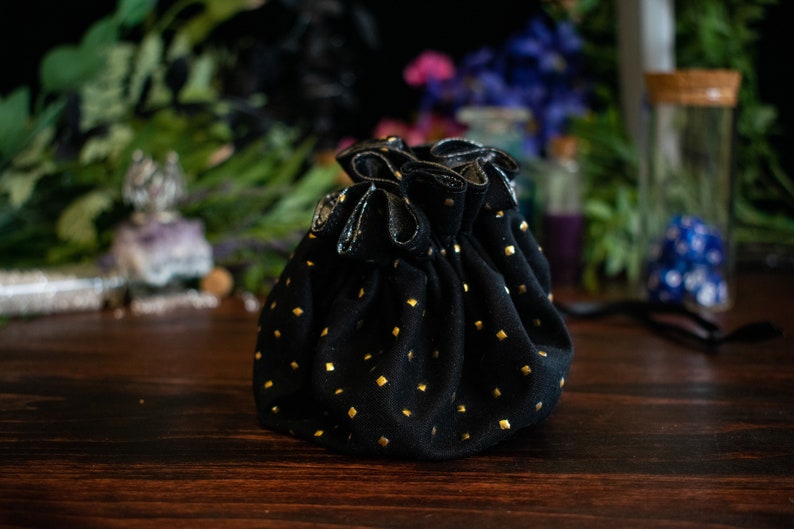 May include: A black drawstring pouch with gold square accents. The pouch is made of a soft material and has a gathered top. The background includes greenery and other decorative items, suggesting a mystical or bohemian theme.