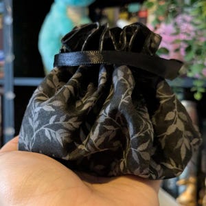 May include: A small, black drawstring pouch with a gray leaf pattern. The pouch is gathered at the top with a black ribbon. The fabric has a matte finish. The pouch is held in a hand.
