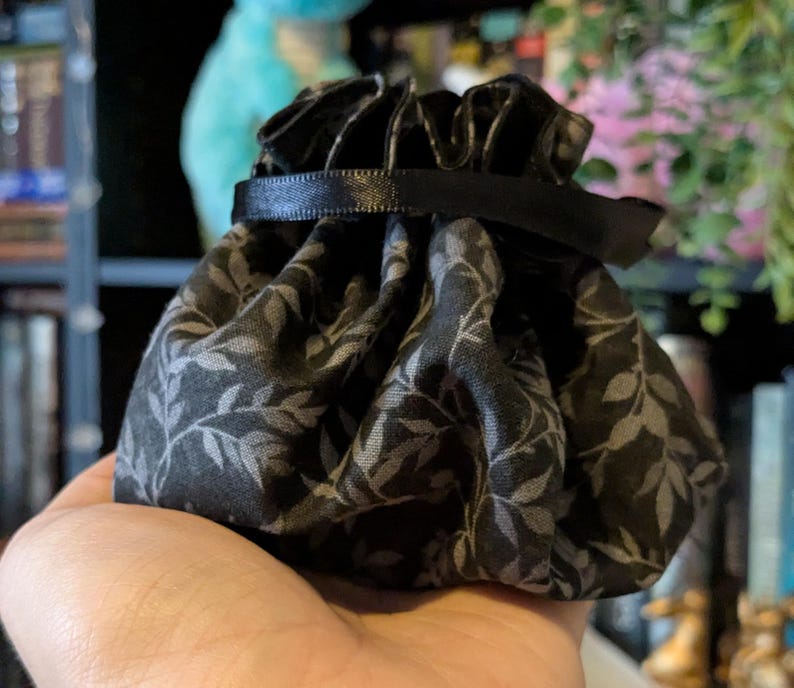 May include: A small, black drawstring pouch with a gray leaf pattern. The pouch is gathered at the top with a black ribbon. The fabric has a matte finish. The pouch is held in a hand.