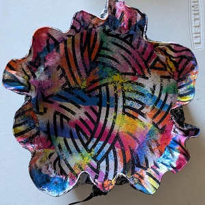 May include: A colorful, abstract bowl with a scalloped edge. The bowl features a vibrant design of black curved lines and multicolored paint splatters in shades of pink, blue, yellow, and orange. The bowl is approximately 10 inches in diameter.