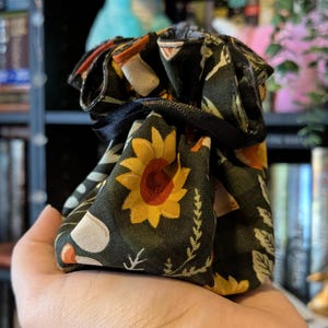 May include: A small drawstring bag with a black background and a pattern of sunflowers, leaves, and other floral elements in yellow, orange, and white. The bag is tied with a black ribbon.
