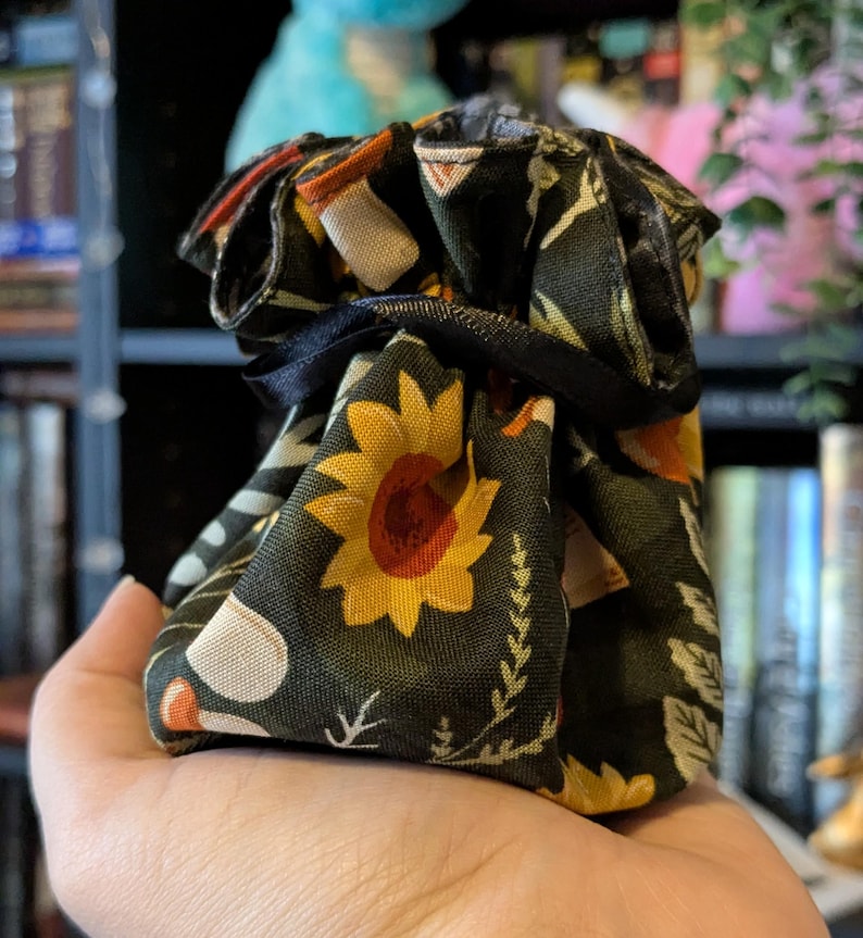 May include: A small drawstring bag with a black background and a pattern of sunflowers, leaves, and other floral elements in yellow, orange, and white. The bag is tied with a black ribbon.