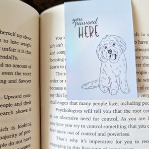 May include: A light blue bookmark with the text "you paused HERE" and a line drawing of a dog. The bookmark is inserted into a book with printed text on the pages.