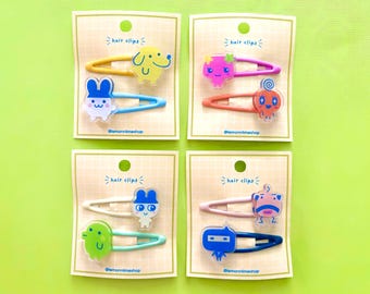 May include: Four sets of hair clips, each with two clips. Each clip features a colorful cartoon character design. The clips are on a cream-colored card with the words "hair clips" and the shop's name. The background is a bright green.