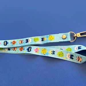 May include: A light blue lanyard with a repeating pattern of colorful cartoon characters. The lanyard has a gold-colored metal clasp and a snap closure. The background is a solid blue.