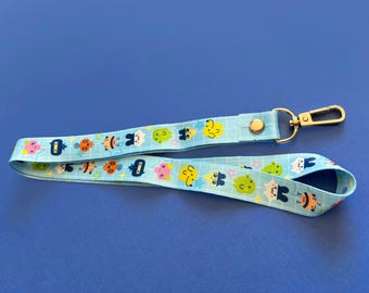 May include: A light blue lanyard with a repeating pattern of colorful cartoon characters. The lanyard has a gold-colored metal clasp and a snap closure. The background is a solid blue.
