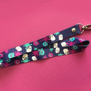 May include: A navy blue lanyard with a repeating pattern of cartoon characters and colorful splatters. The lanyard has a metal clasp and a snap closure. The background is a solid pink color.