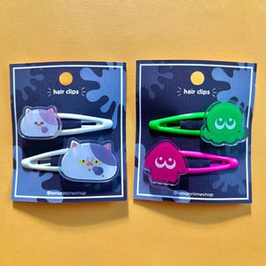 May include: Two sets of hair clips on a yellow surface. One set features cat designs with white and purple accents, the other set has green and pink squid designs. Each set is on a blue card with the text "hair clips".