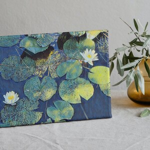 Op de afbeelding: Een canvas print van een blauwe vijver met witte waterlelies en groene waterleliebladeren. De print is ingelijst en leunt tegen een wit oppervlak.