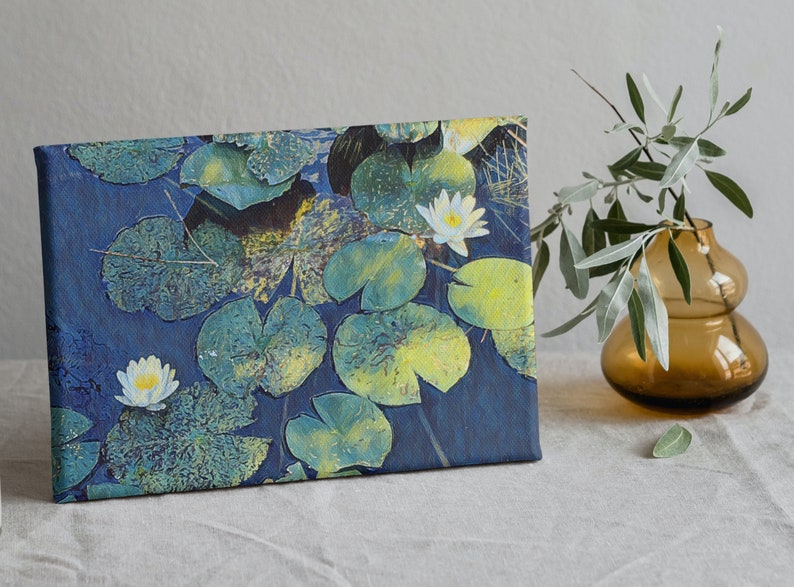 Op de afbeelding: Een canvas print van een blauwe vijver met witte waterlelies en groene waterleliebladeren. De print is ingelijst en leunt tegen een wit oppervlak.
