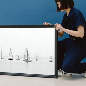 K&ouml;nnte beinhalten: Ein schwarzer gerahmter Druck einer Bleistiftzeichnung von Segelbooten auf dem Wasser. Der Druck wird von einer Person in einem dunkelblauen Overall gehalten.