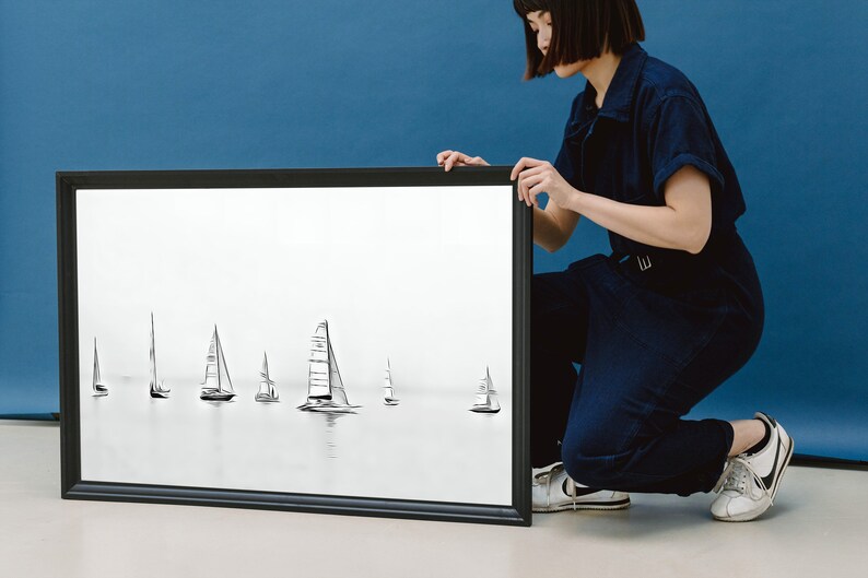 K&ouml;nnte beinhalten: Ein schwarzer gerahmter Druck einer Bleistiftzeichnung von Segelbooten auf dem Wasser. Der Druck wird von einer Person in einem dunkelblauen Overall gehalten.