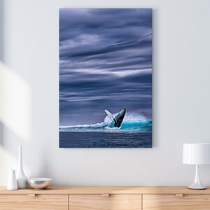 May include: A canvas print depicts a whale breaching from a turquoise wave under a dramatic, cloudy sky. The artwork is displayed on a white wall above a wooden cabinet with decorative items.
