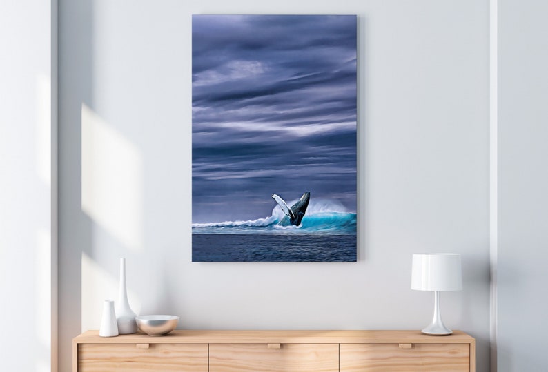 May include: A canvas print depicts a whale breaching from a turquoise wave under a dramatic, cloudy sky. The artwork is displayed on a white wall above a wooden cabinet with decorative items.
