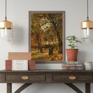 K&ouml;nnte beinhalten: Ein gerahmtes Gem&auml;lde, das eine Waldszene mit Personen zeigt, die auf einem Weg gehen. Das Gem&auml;lde h&auml;ngt an einer Wand &uuml;ber einem Holz-Konsolentisch mit zwei Schubladen. Zwei goldene Pendelleuchten h&auml;ngen &uuml;ber dem Tisch, und eine Topfpflanze und B&uuml;cher stehen auf dem Tisch.