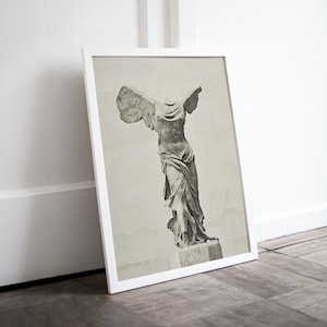 May include: A black and white photograph of the Winged Victory of Samothrace statue, a famous Greek sculpture. The statue is displayed in a white frame.